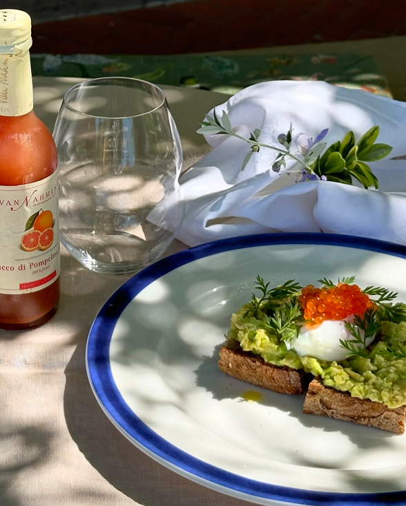 Tavola apparecchiata all'aperto ripresa in diagonale con avocado toast e spremuta d'arancia.
