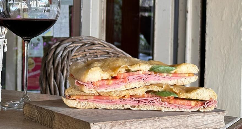 Due schiacciate farcite di salumi, verdura e salse appoggiate su un tagliere con accanto un calice di vino rosso.