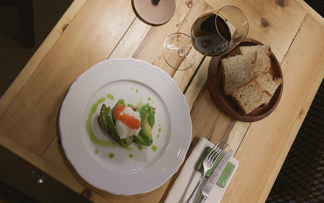 Tavola vista dall'alto con piatto avocado toast, calice di vino rosso e cesto del pane fatto in casa.